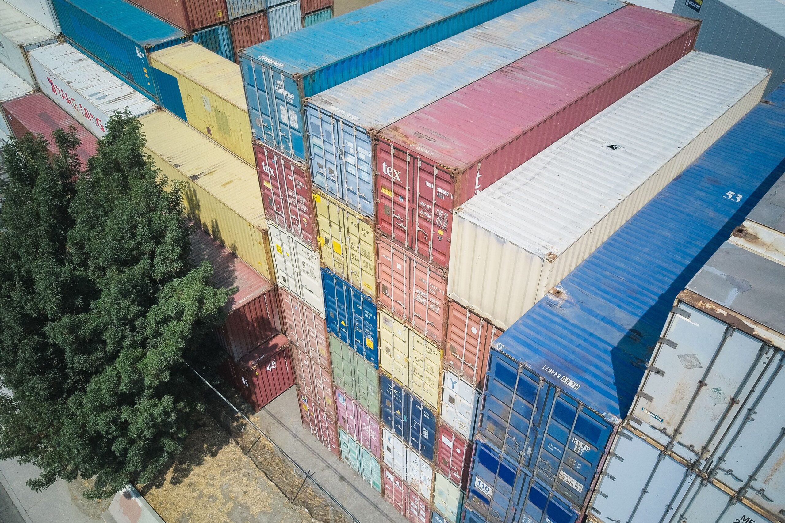 An aerial shot of multi-colored stacked shipping containers near a tree.